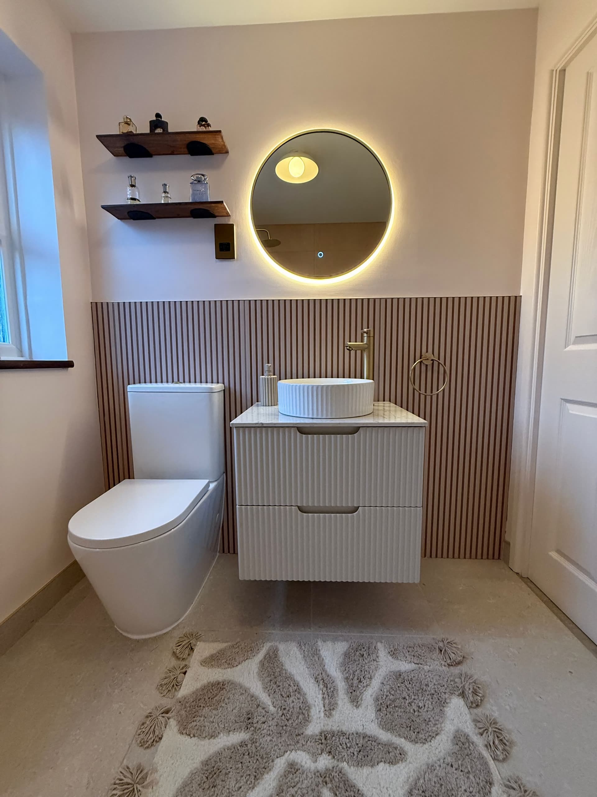 Finished bathroom with slatted timber panelling, a round backlit mirror and a fluted wall-hung vanity with a brass tap