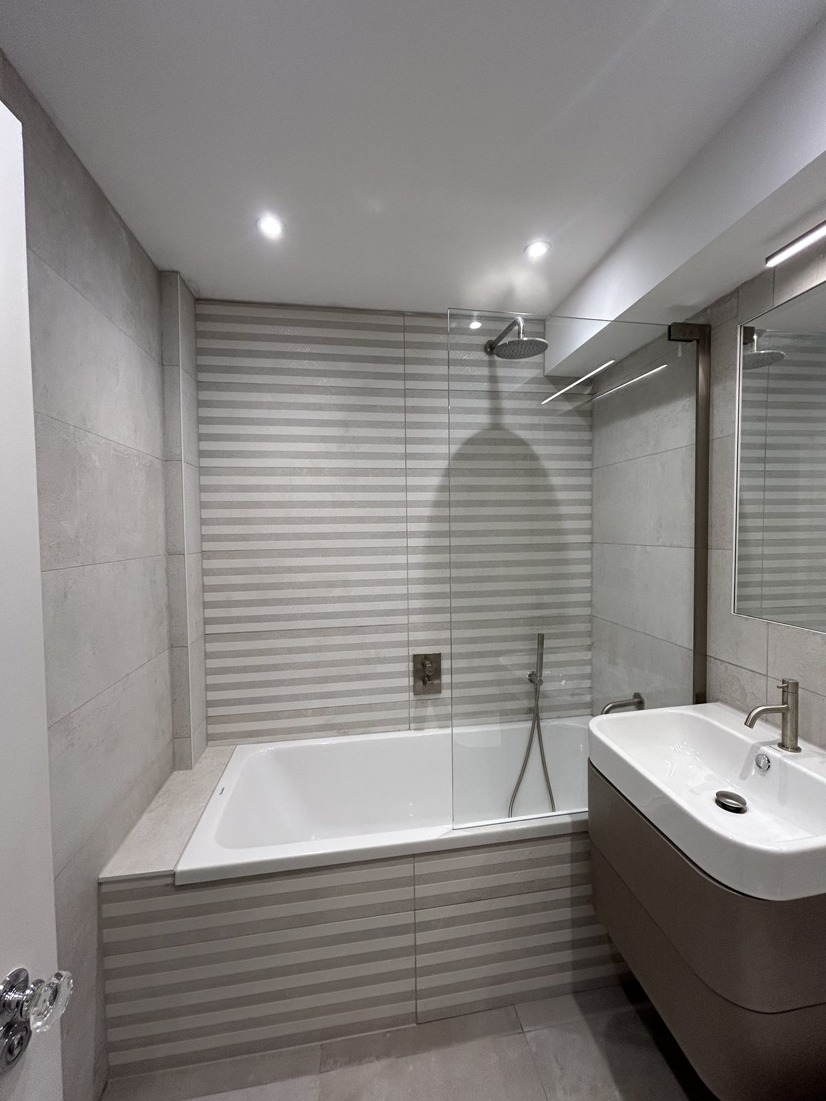 Bath with a rainfall shower over behind a glass screen, against a striped relief tile wall