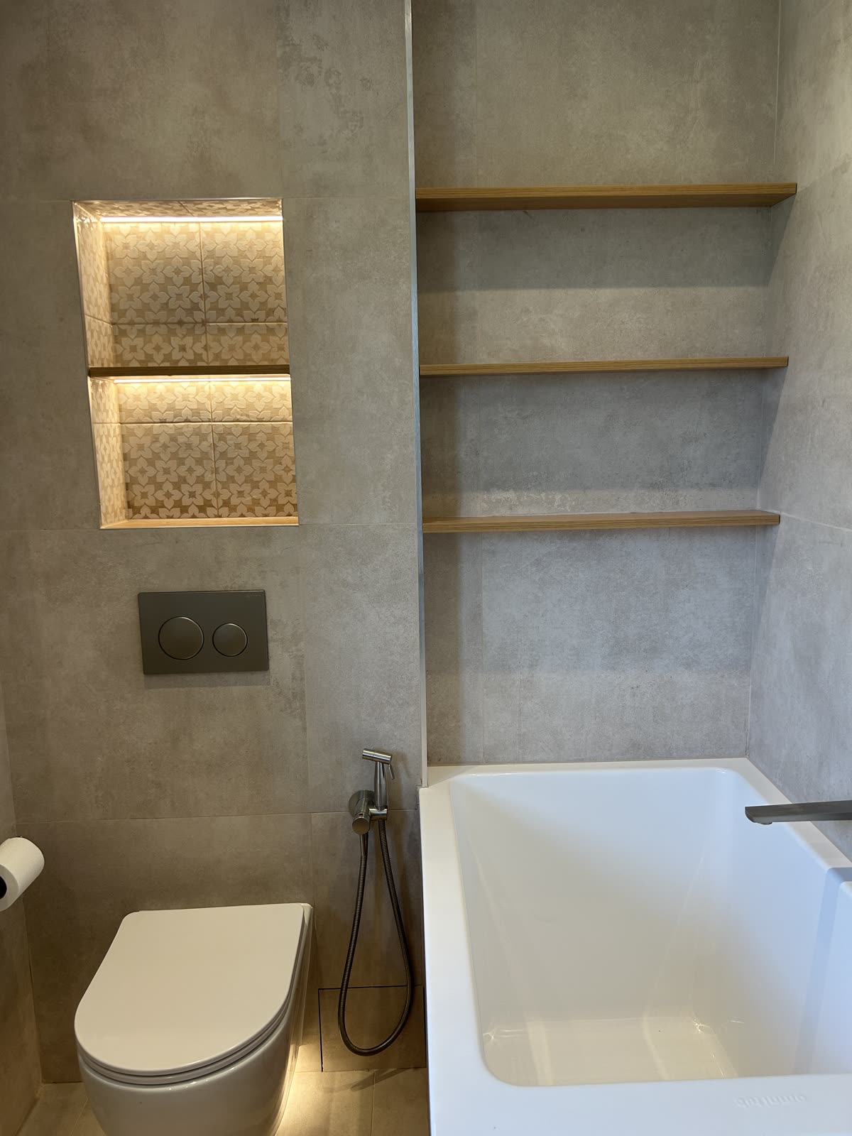 Wall-hung WC beside a backlit niche, with oak shelving above the bath