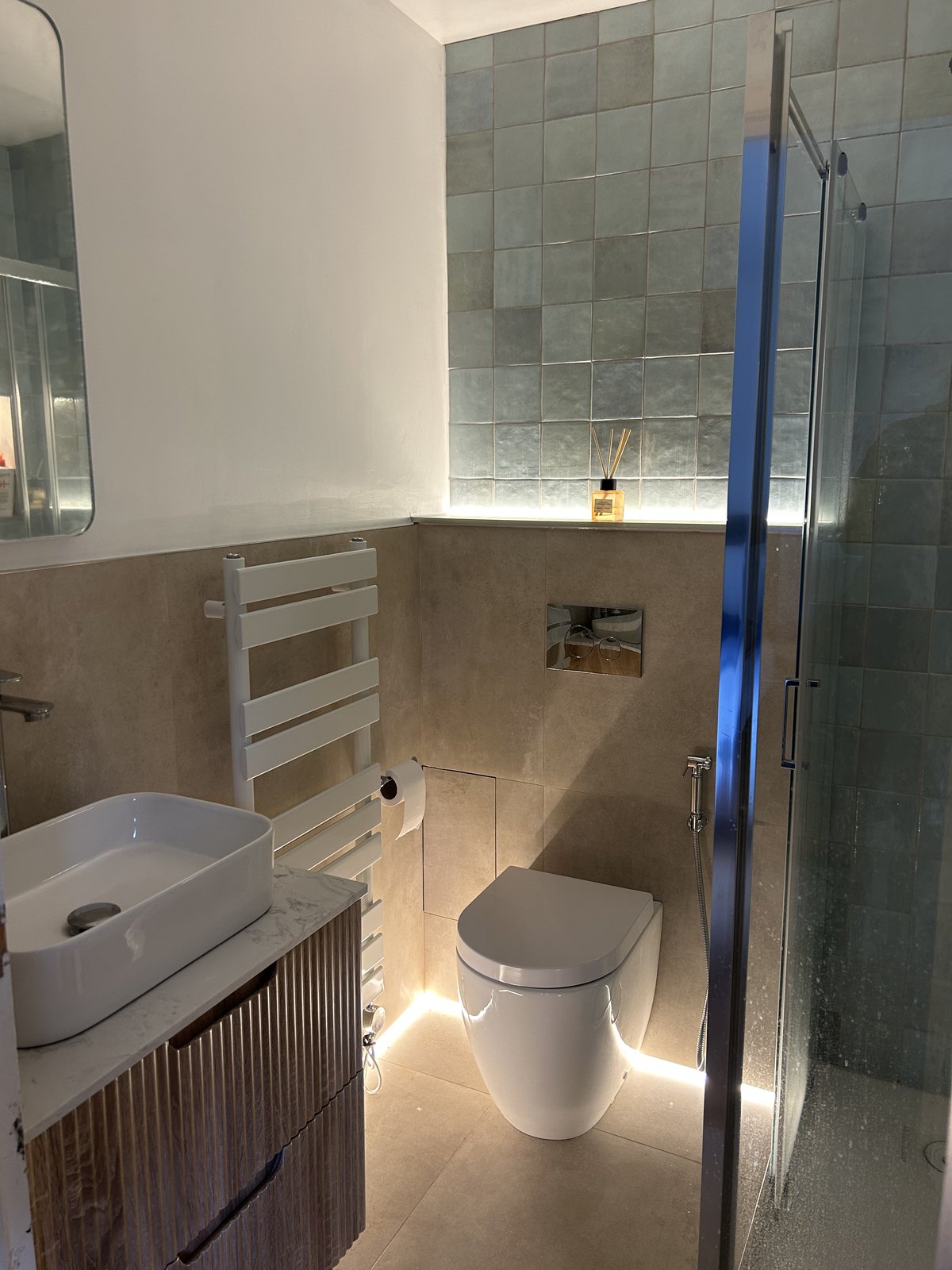 Bathroom with mottled green glazed tile above stone-effect porcelain, a fluted vanity and a lit ledge