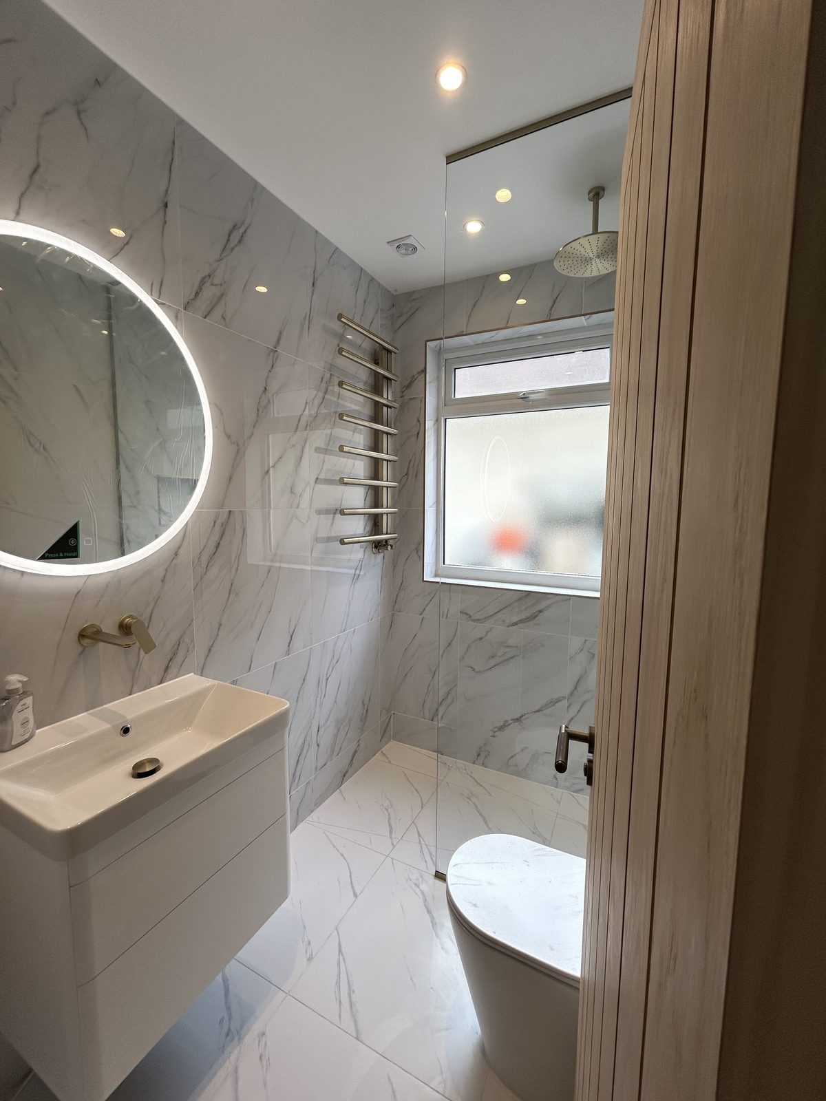 Marble-effect bathroom with a round backlit mirror, brass ladder radiator and walk-in shower
